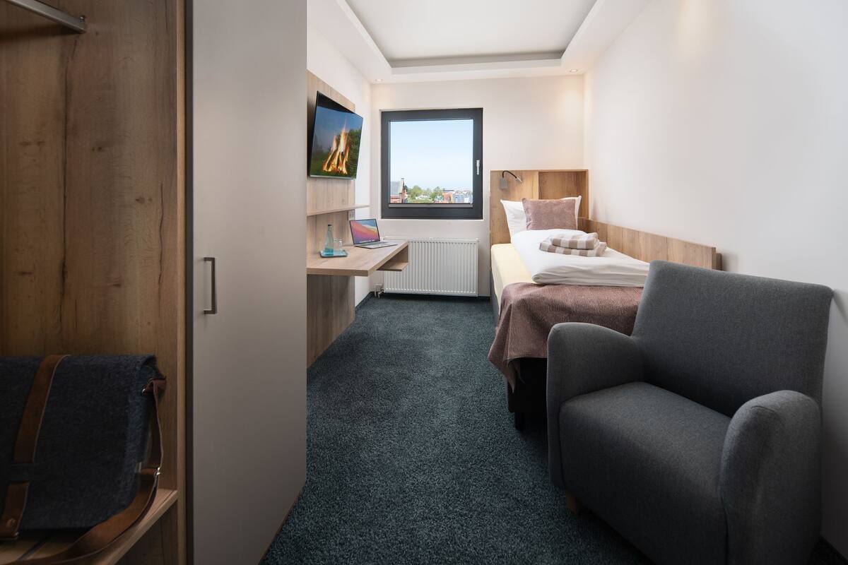 Ein kleines Hotelzimmer mit Bett, Schreibtisch, Stuhl und Blick auf die Stadt durch ein Fenster.
