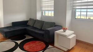 Een moderne woonkamer met een donkergrijze modulaire bank, een witte bijzettafel met een vaas, en een groot raam met zonwering.
