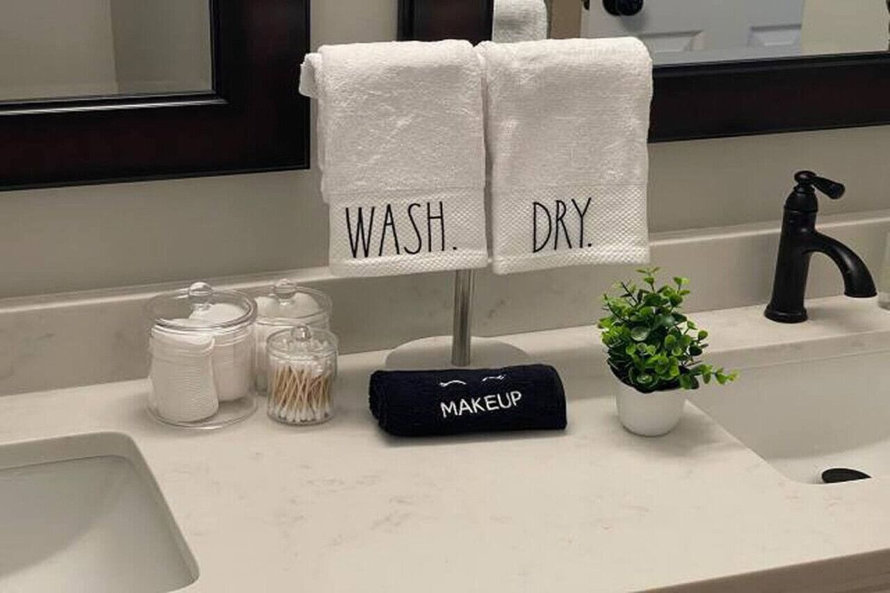 A bathroom with two towels labeled 'WASH.' and 'DRY.' hanging on a rack, a makeup bag, and a small potted plant on the sink counter.