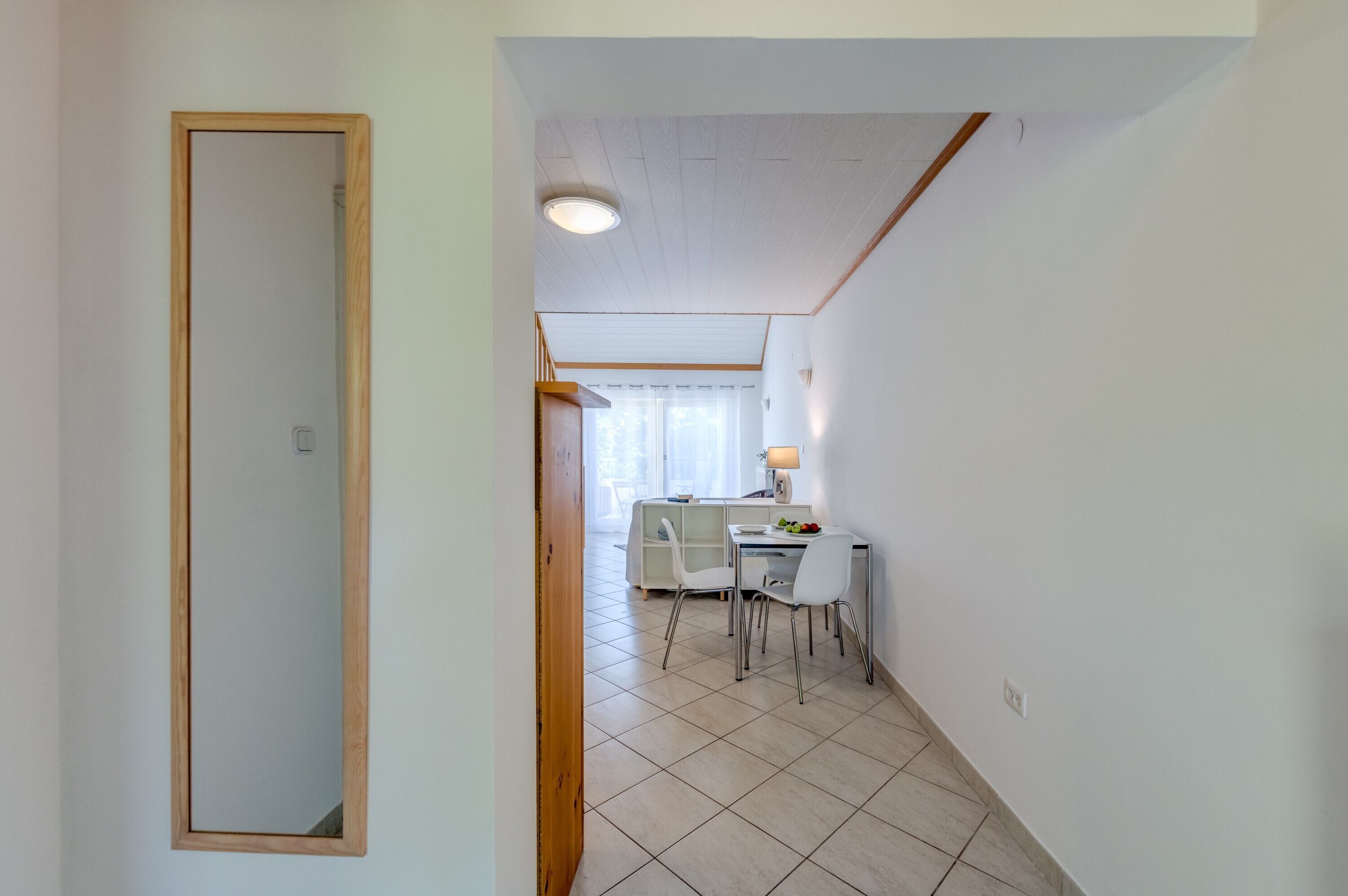 Un pasillo que conduce a un comedor con una mesa y sillas blancas, una lámpara y un ventanal con cortinas.