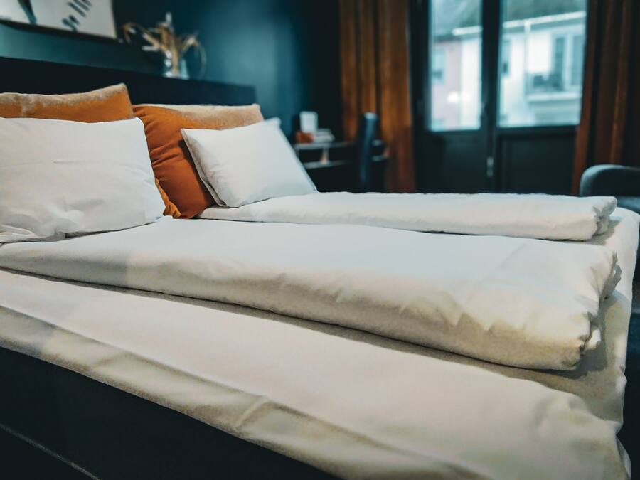 Ein Doppelbett mit weißen und orangefarbenen Kissen, ein dunkles Kopfstück und Blick auf die Straße durch ein Fenster.