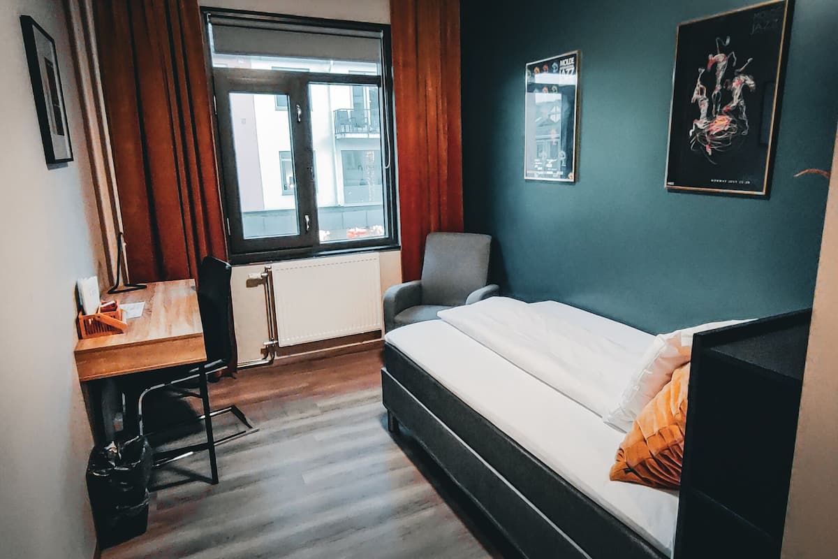 A bedroom with a bed, a desk, and a chair. There is a window with curtains, a radiator, and a poster on the wall.