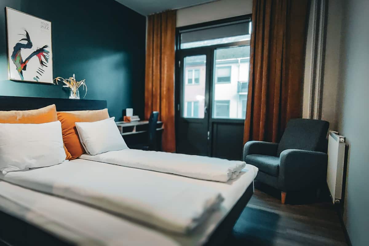 A hotel room with a bed, a chair, a desk, a lamp, and a window with curtains.