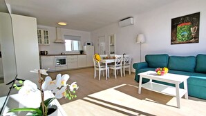 Ein modernes Wohnzimmer mit einem türkisfarbenen Sofa, einem weißen Couchtisch und einem Essbereich mit weißen Stühlen. Im Hintergrund ist eine Küche mit weißen Schränken und einer Mikrowelle zu sehen.