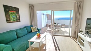 Ein modernes Wohnzimmer mit einem türkisfarbenen Sofa, einem kleinen weißen Tisch mit Obstschale, einem Fernseher und einem Balkon mit Meerblick.