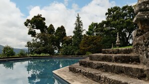 Una piscina in pietra con gradini che scendono verso l'acqua, circondata da alberi e un cielo sereno.