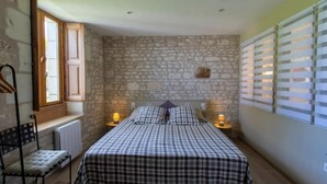 Ein Schlafzimmer mit Steinwand, einem Bett mit karierter Bettwäsche, einem Stuhl und einem Fenster mit Jalousien.