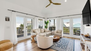 Un salon spacieux avec un grand canapé, un fauteuil rond et une table basse. Un tapis à motifs recouvre le sol, et la pièce est dotée de grandes fenêtres offrant une vue sur l’extérieur.