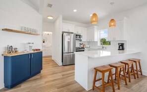 A modern kitchen with white cabinetry, a stainless steel refrigerator, wooden stools, and pendant lighting.
