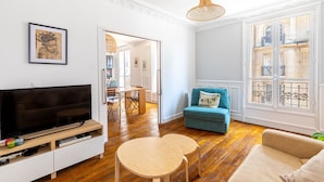 Una sala de estar con televisor, sofá, una mesita y una silla. Se ve una ventana con vistas a edificios.