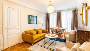 Un salon bien éclairé, avec un canapé de couleur moutarde, une table basse ronde et un coin repas comprenant une table et des chaises.