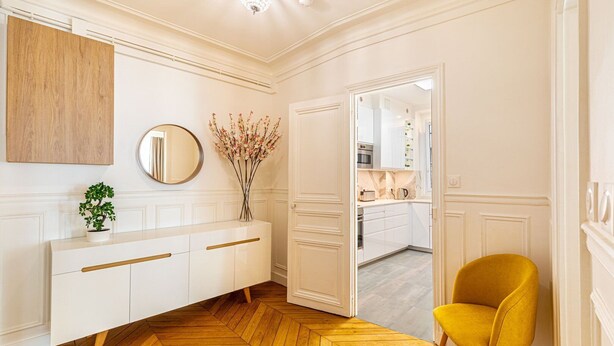Une salle de bain moderne avec un parquet en bois disposé en motif chevrons, un meuble-lavabo blanc avec des poignées dorées, un miroir, un vase avec des fleurs et une chaise jaune.