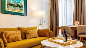 Un salon avec un canapé de couleur moutarde, une table basse ronde sur laquelle se trouvent une bouteille et des verres, une lampe, et un coin repas avec des chaises et une table.