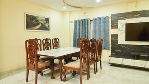 A dining area with a marble table and wooden chairs, a painting on the wall, and a TV on a marble cabinet. - Bliss Farm Stay-IRIS (Hyderabad)