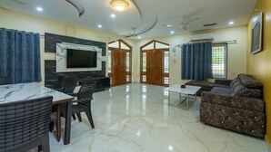 A spacious living room with a marble floor, a flat-screen TV mounted on the wall, a sofa, a coffee table, and a dining area with chairs. - Bliss Farm Stay-IRIS (Hyderabad)