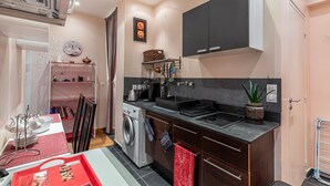 A compact kitchen with a washing machine, a stove, and a sink. There is a dining area with a table and chairs, and a shelf with kitchen utensils. - GuestReady - Minimalist stay near Opéra Bastille (Paris)
