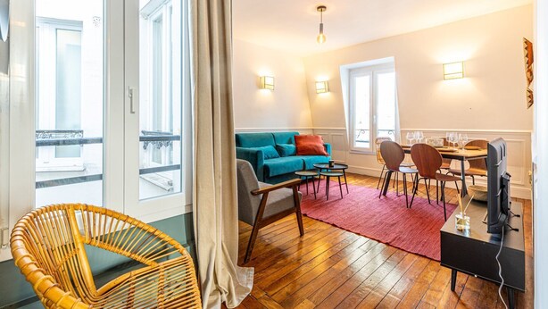 Een moderne woonkamer met een blauwgroene bank, houten vloeren, een eethoek met stoelen en een tafel, en een televisie.