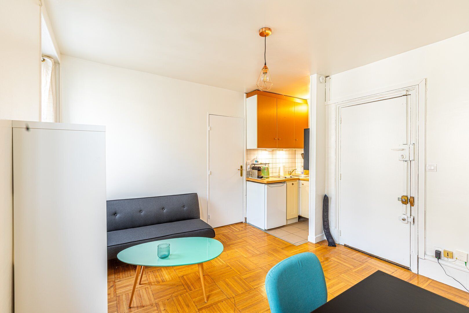 A compact living space with a sofa, a small round table, a chair, a kitchenette, and a door.