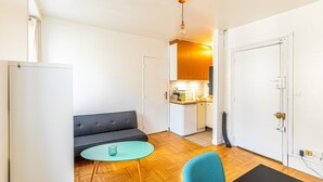 Un espace de vie compact comprenant un canapé, une petite table ronde, une chaise, un coin cuisine et une porte.