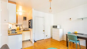 Une cuisine compacte équipée d’un four à micro-ondes, d’un petit réfrigérateur et d’un plan de travail en bois. On y trouve une chaise de couleur turquoise et une petite table sur laquelle est posé un objet jaune.
