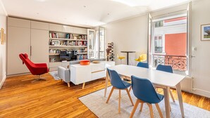 Un salon moderne avec un canapé blanc, un fauteuil rouge et des chaises bleues disposées autour d’une table blanche. On peut voir une bibliothèque remplie de livres et un téléviseur.
