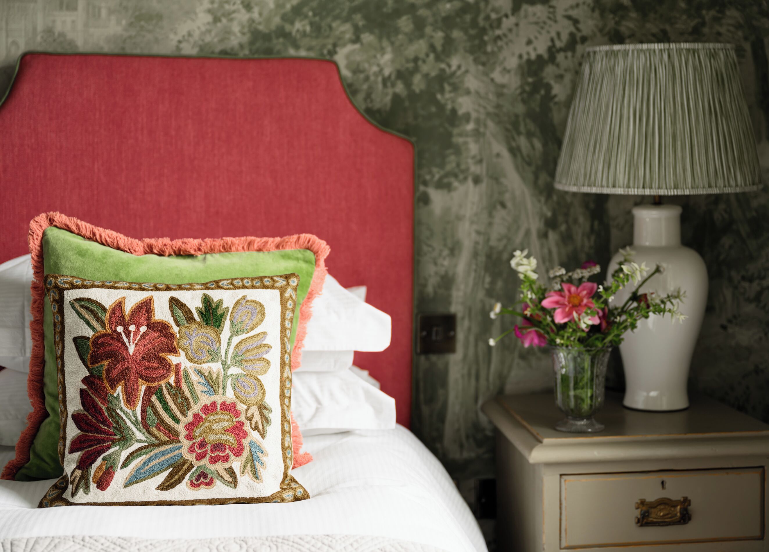 A neatly made bed with a red headboard, a decorative pillow, a bedside table with a lamp and a vase of flowers.
