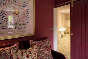 Una habitación con una pared roja, una obra de arte floral enmarcada, un sofá rojo con cojines estampados y vistas a un baño con una bañera blanca y un inodoro.