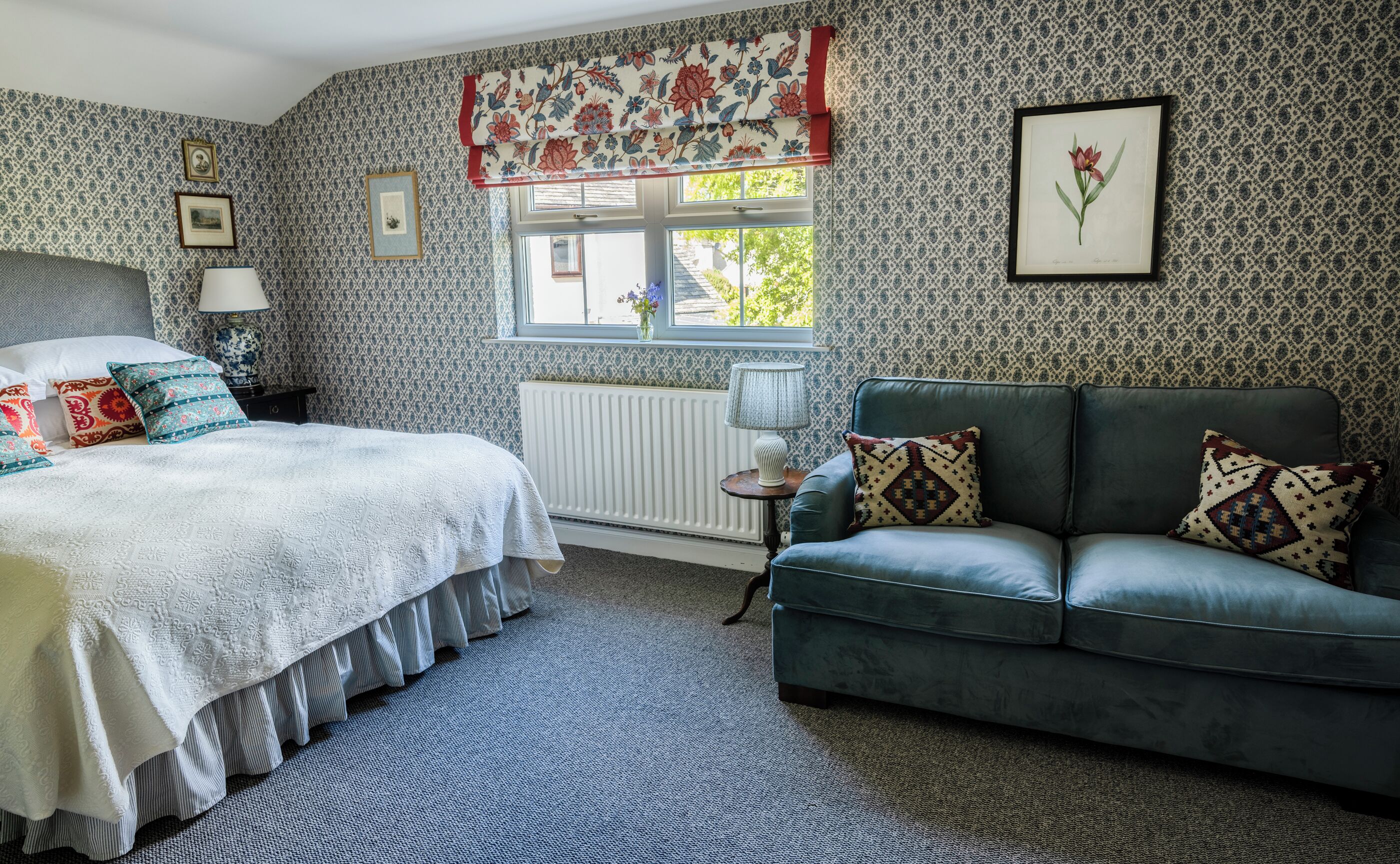 Un dormitorio con cama, un sofá verde, papel tapiz estampado, una ventana con un adorno floral y una ilustración botánica enmarcada.