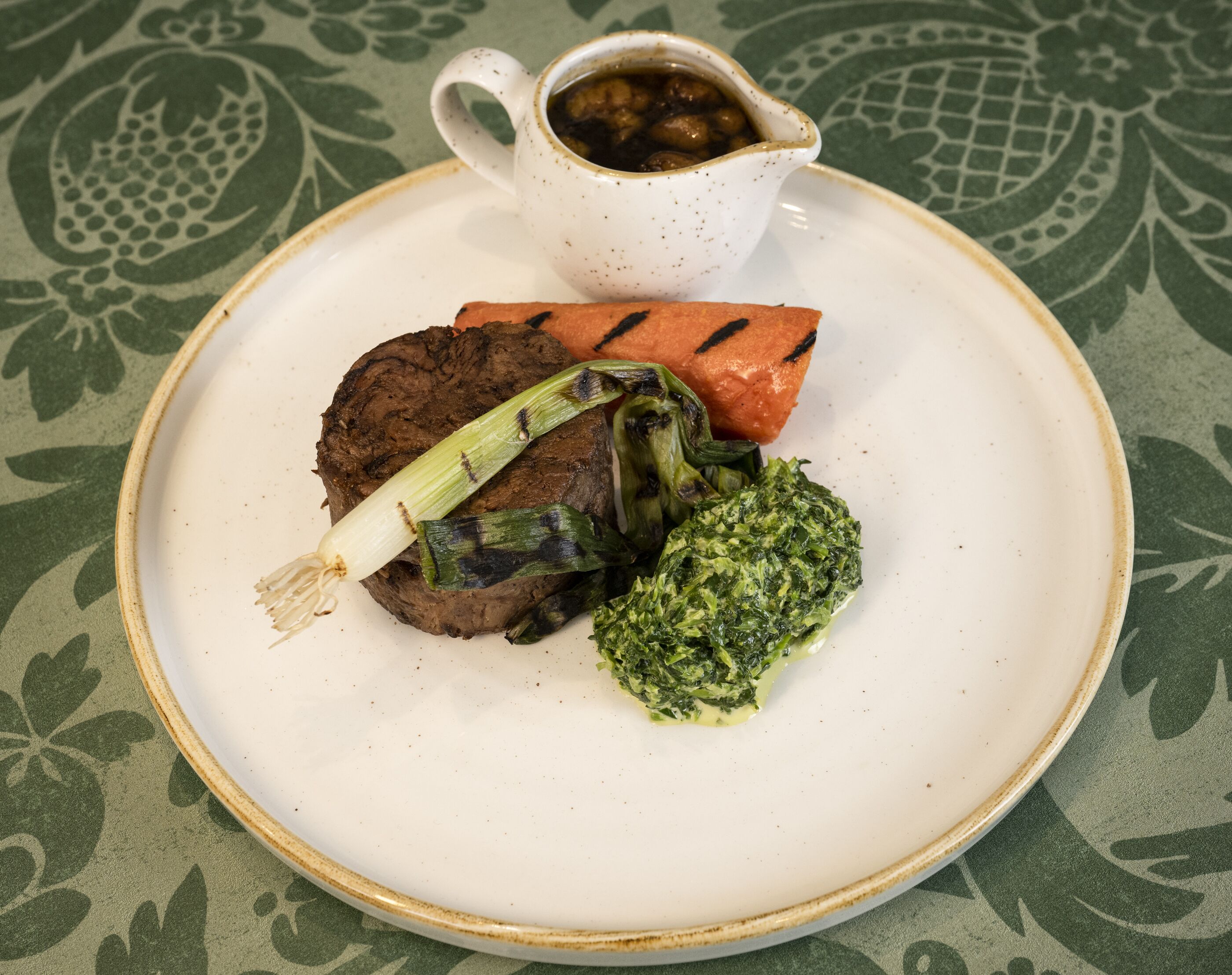 A plate with a steak, grilled vegetables, and a cup of sauce.