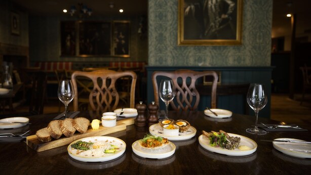 A dining table set with plates of food, wine glasses, and cutlery. There are chairs around the table, and a painting is visible on the wall. - George and Dragon Clifton (Penrith)