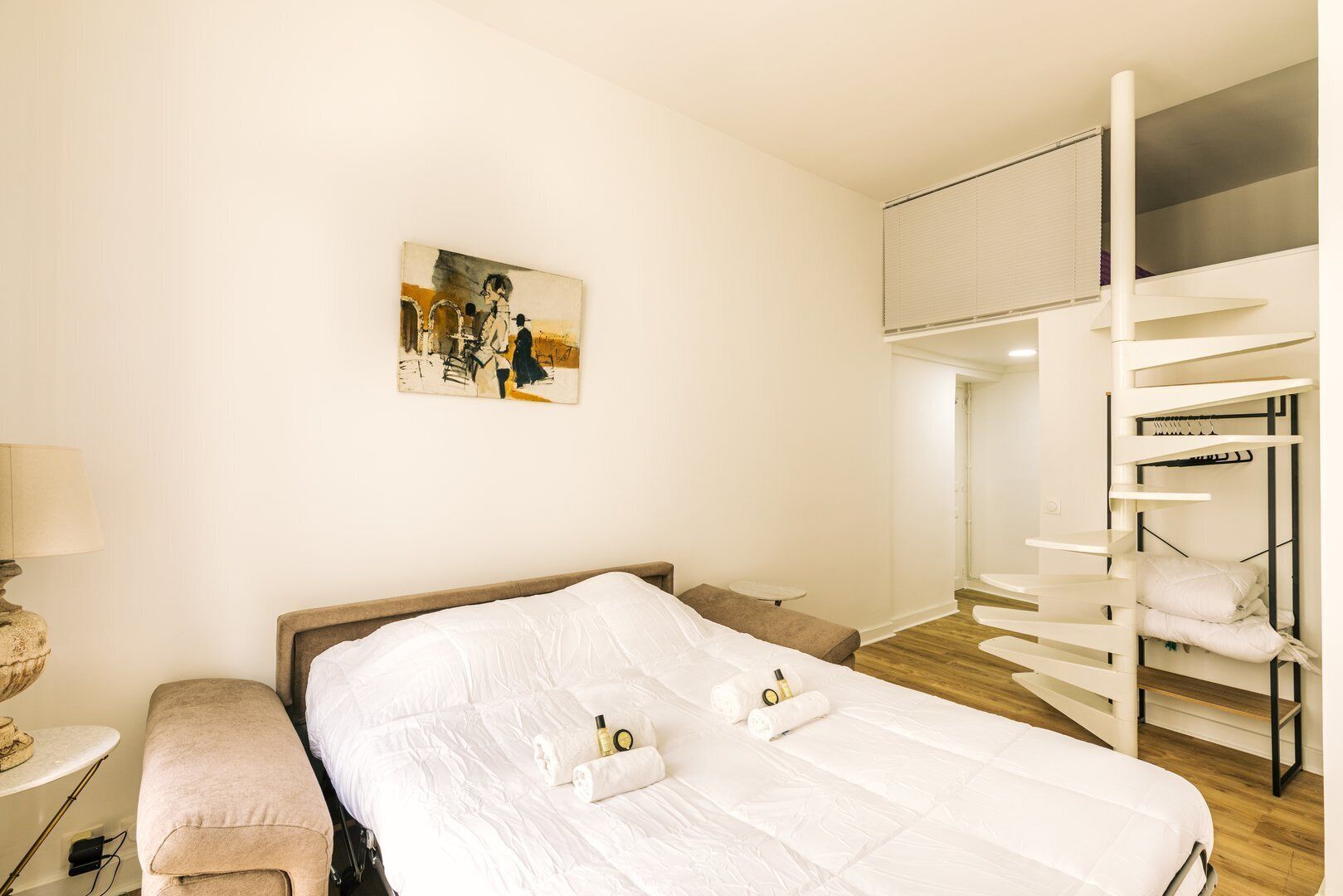 Habitación de hotel con una cama grande, una mesita de noche pequeña, un banco, un cuadro en la pared y una litera en la esquina.