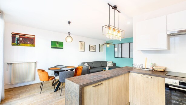 Una cocina moderna con una isla central, armarios de madera y un horno integrado. Hay una zona de comedor con una mesa redonda y sillas, y un sofá en la sala de estar.