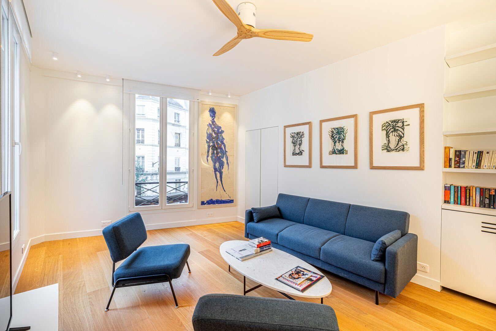 A modern living room with a blue sofa, two armchairs, a coffee table with magazines, and a ceiling fan. There are framed artworks on the wall and a bookshelf filled with books.