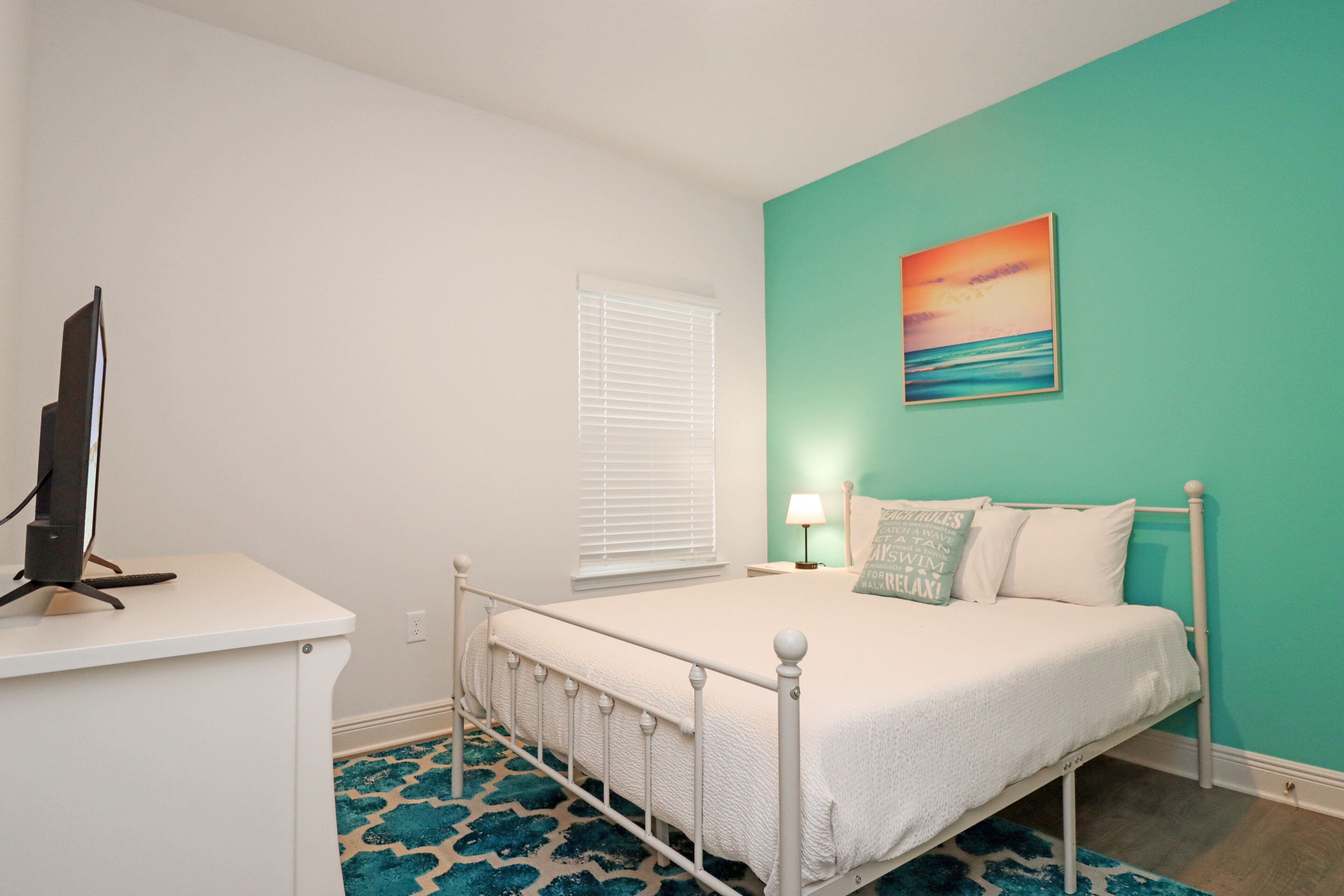 A bedroom with a bed, a desk, a computer, a window with blinds, a painting, and a patterned rug.