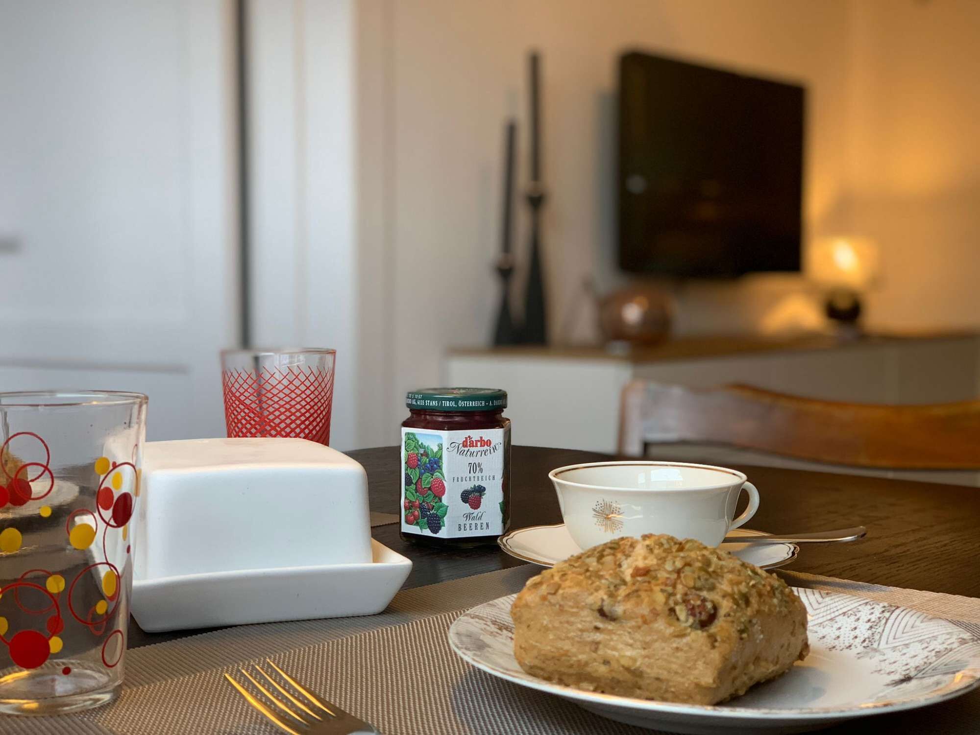 Uma mesa com um frasco de Darbo Naturleber, um copo com padrão vermelho, uma xícara branca e um prato com uma bolacha.