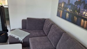 Un salon moderne avec un canapé d’angle gris, une table d’appoint blanche et un grand tableau représentant un paysage urbain accroché au mur.