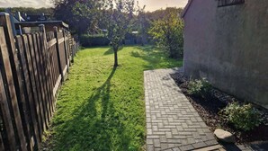 Un jardin avec un chemin dallé, une clôture en bois et un arbre qui projette une ombre.