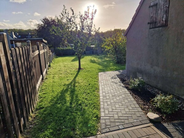 Un patio trasero con un sendero pavimentado, cerca de madera y un árbol proyectando una sombra.