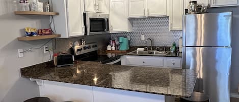 Una cocina moderna con armarios blancos, encimera de granito, refrigerador de acero inoxidable y cuatro taburetes.