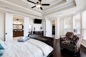 A bedroom with a large bed, a ceiling fan, a TV mounted on the wall, and a view of the kitchen through an open door.