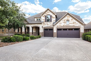 A two-story stone house with a garage, a large tree, and well-maintained landscaping.