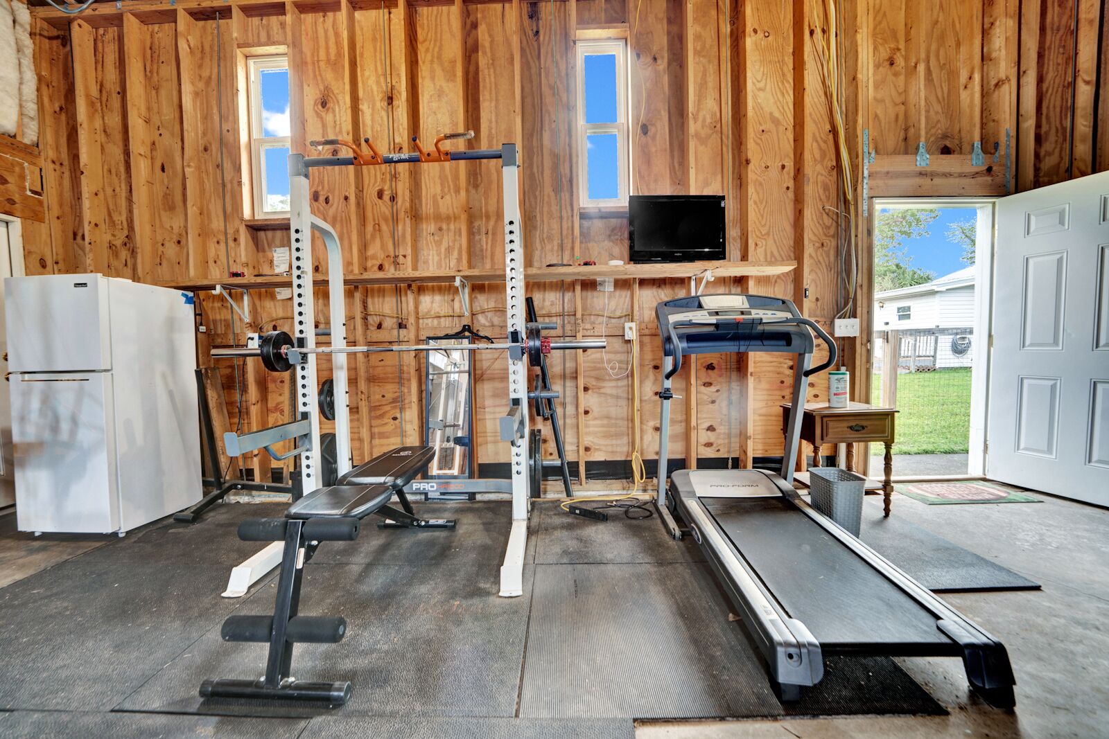 A home gym with a bench press, squat rack, treadmill, and a TV mounted on the wall.