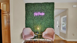 Una habitación con una pared verde cubierta de follaje, dos sillas rosadas, una pequeña mesa redonda y un letrero de neón que dice 'Palm Paradise'.