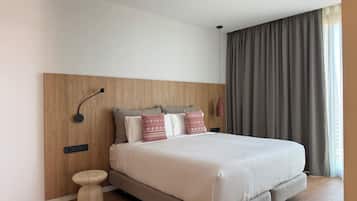 Una habitación de hotel moderna con una cama grande, un cabecero de madera, una mesita de noche y un ventanal con cortinas.