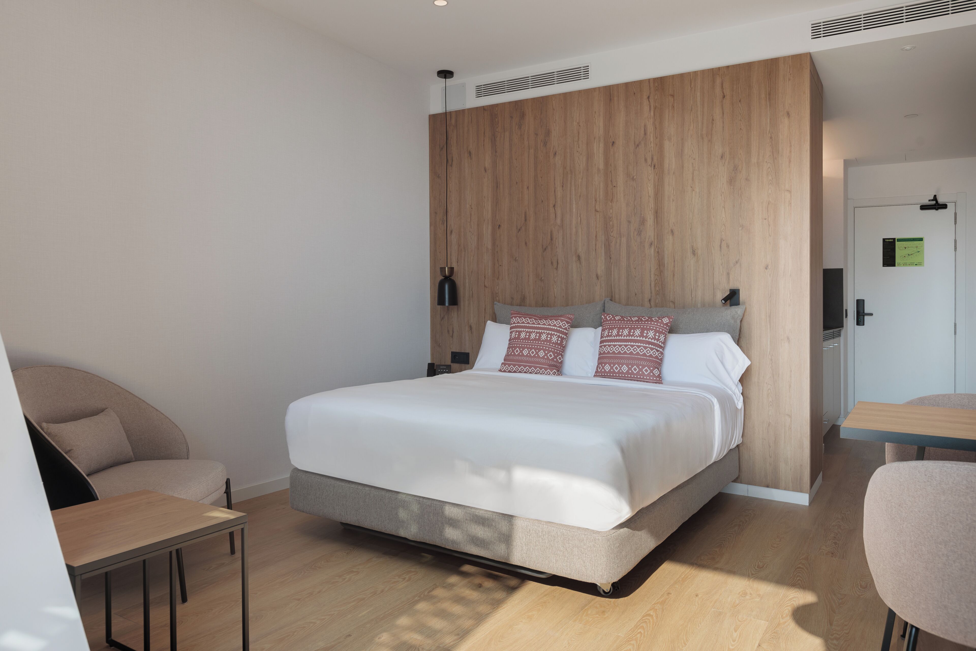 Una habitación de hotel moderna con un cabecero de madera, una cama con almohadas estampadas, una mesita pequeña y una silla.