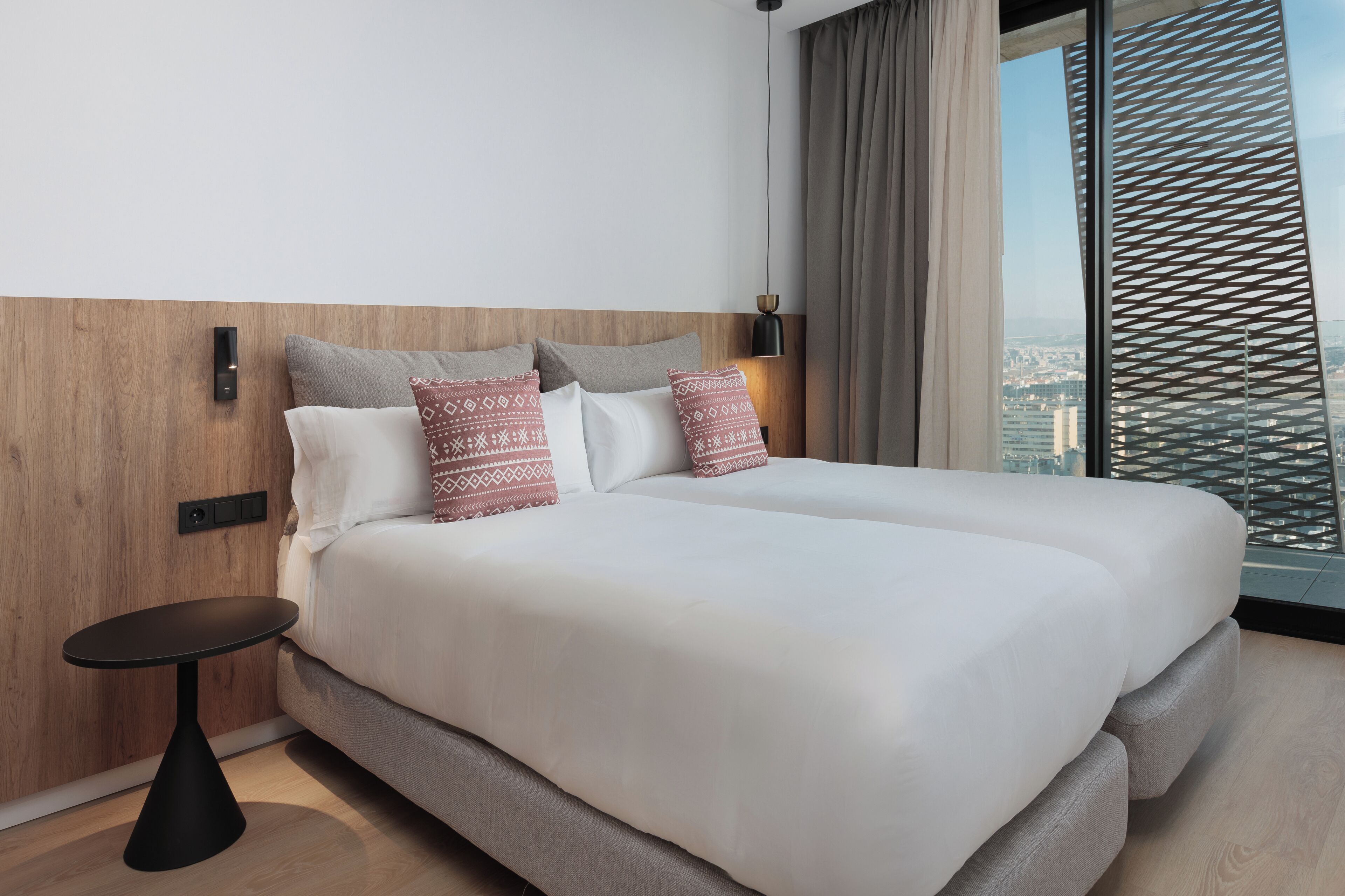 Habitación de hotel moderna con una cama grande, una mesita auxiliar y vistas a la ciudad desde la ventana.