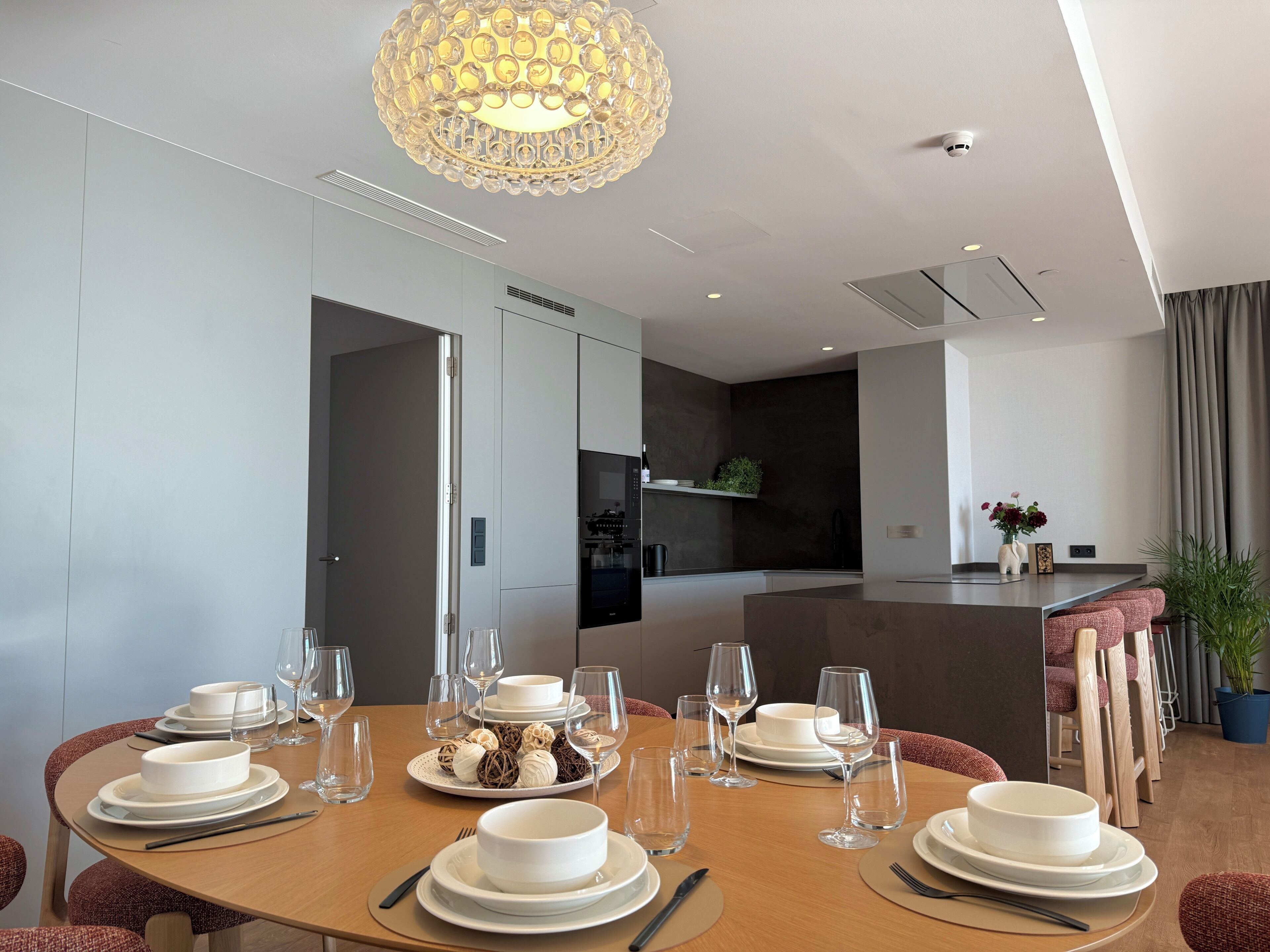 Een moderne eetruimte met een ronde tafel gedekt voor een maaltijd, een keuken op de achtergrond en een decoratieve kroonluchter.