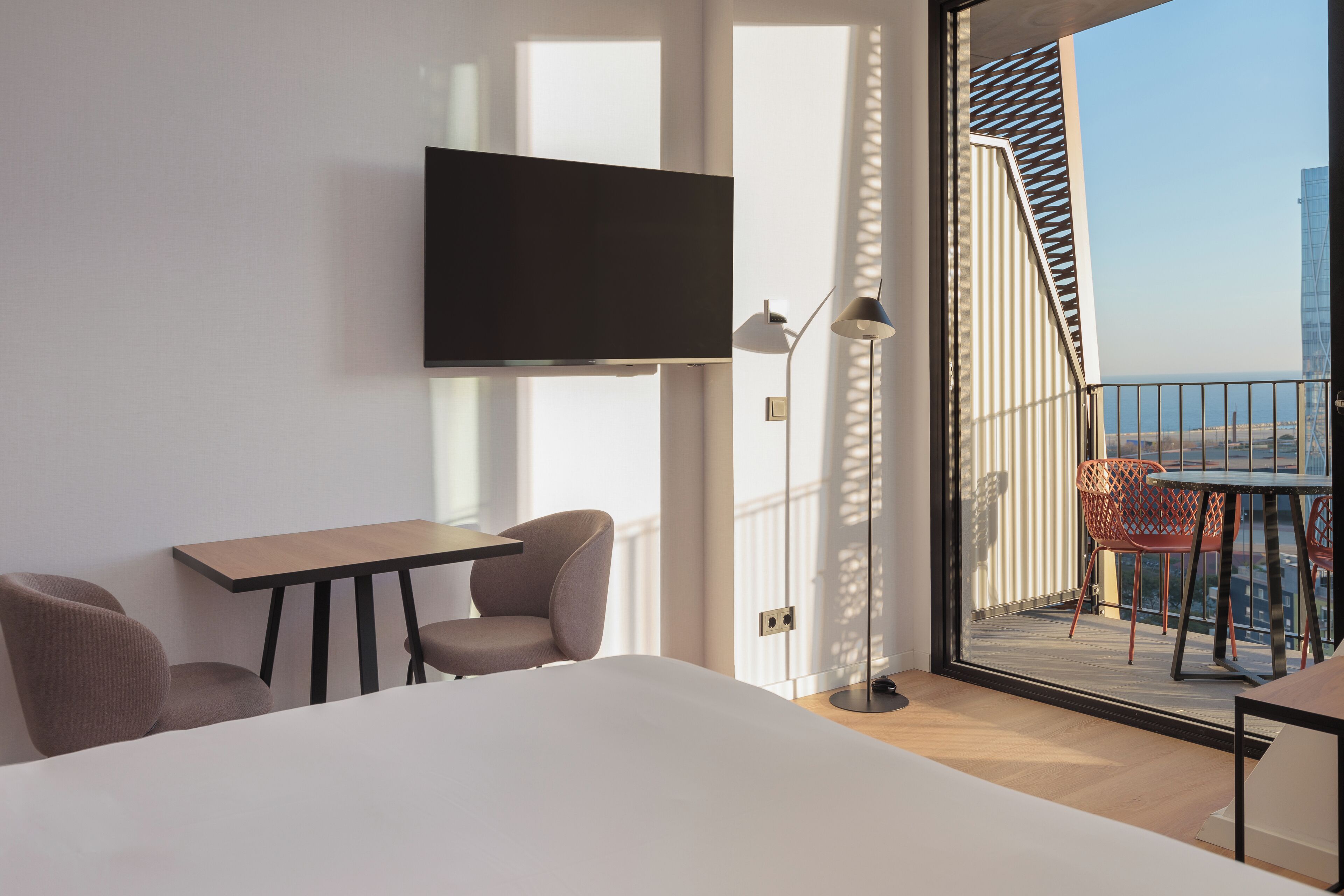 Habitación de hotel moderna con cama, escritorio, dos sillas, televisor montado en la pared y balcón con vista a la ciudad.