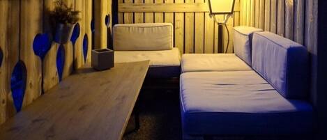 Un rincón exterior acogedor con una mesa de madera y un banco acolchado, iluminado por una lámpara colgante.