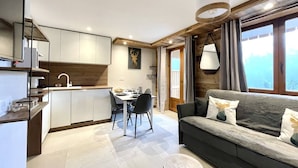 A modern living room with a grey sofa, a dining table with chairs, a kitchen area with white cabinets, and a large window with curtains. - N°12 : 4 people flat with balcony, incl. swimmingpool, sauna, hammam (CHAMPAGNY EN VANOISE)